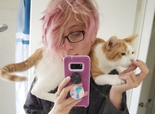 A young woman with short pink and blonde hair wears black rimmed glasses as she poses for a mirror selfie while an orange and white cat lies draped around her shoulders.