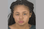A young woman with long dark brown braided hair worn half pulled back has one eye half closed as she stares at the camera during her mugshot.