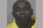 A middle-aged bald man frowns and leans to the side as he wears a yellow jumpsuit during his mugshot.