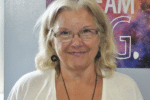 A smiling middle-aged white woman with glasses and long, light-colored hair wearing a white top, standing in front of a colorful poster.