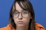 A young white woman with long brown hair wearing glasses and an orange top, looking into the distance with a neutral expression, against a blue background.