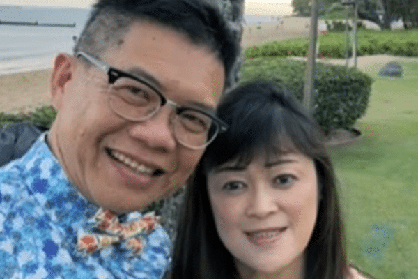 A smiling man and woman pose together outdoors near a beach, standing on a grassy area with greenery and the shoreline in the background.