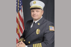 A man in a dark formal fire chief uniform poses in front of an American flag, wearing a white cap with gold trim and a badge on his chest.