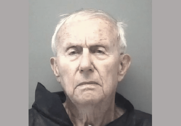 Elderly man with short white hair wearing a dark garment, photographed against a plain background in a mugshot-style image.