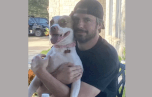 A man in a black shirt and backwards cap hugs a happy, brown-and-white dog wearing a pink collar as they sit outdoors near a stone building.