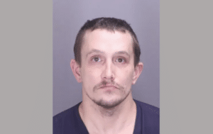 A mugshot of a man with short dark hair and slight facial hair, wearing a dark shirt against a plain gray background.