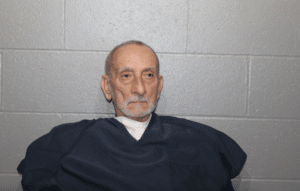 An elderly man with short gray hair and a trimmed beard, wearing a dark blue prison uniform, sits against a gray cinder block wall.