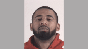 A man with short hair and a beard, wearing a red hoodie, looks directly at the camera against a neutral background.