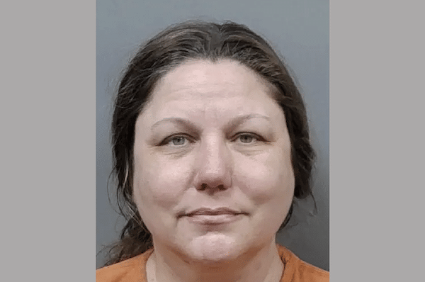 A middle-aged woman with shoulder-length brown hair and a neutral expression, photographed against a plain gray background while wearing an orange shirt.