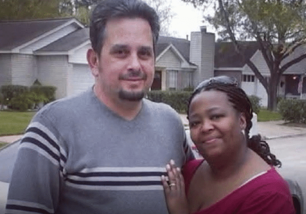 A man and woman standing closely together outside in front of suburban houses, smiling gently for the camera.