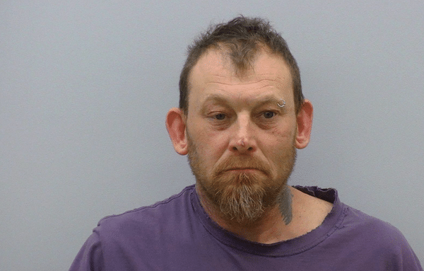 A middle-aged man with short, dark hair and a beard wearing a purple shirt, photographed in front of a plain light-colored background, facing the camera with a neutral expression.