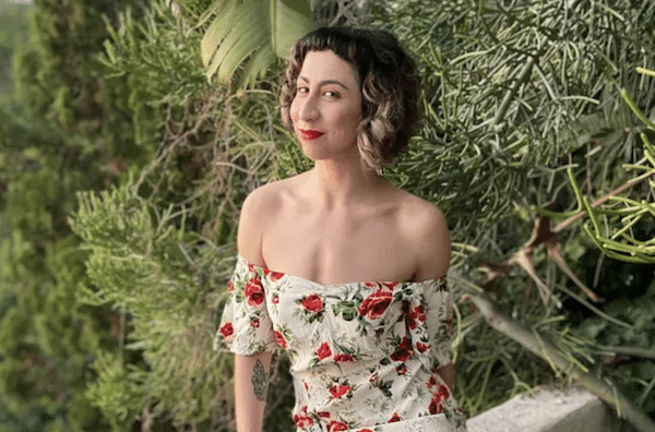 A woman with short, curly hair wearing a white, off-the-shoulder floral dress stands outside in front of tall green bushes, smiling with red lipstick.