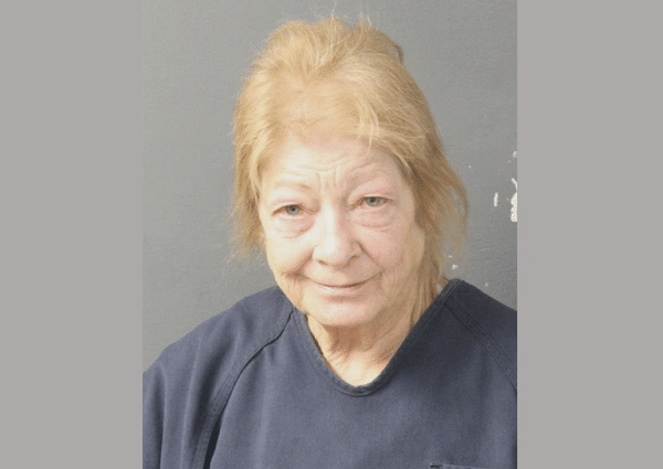 A woman with light-colored hair and a blue top stands against a gray background, looking directly at the camera with a slight smile.