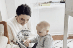 A woman with dark hair and a white sweatshirt sits on the floor next to a young child, who is playing and looking down, with soft daylight coming through a window in the background.
