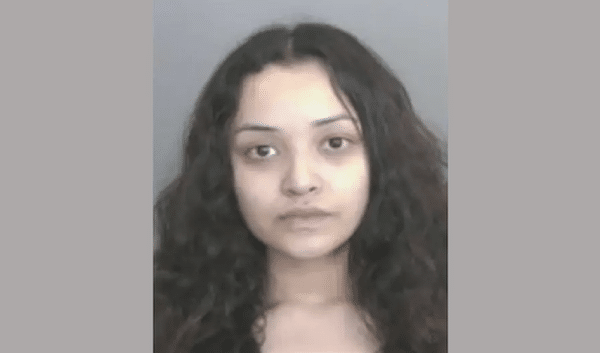 A young woman with long, curly dark hair stares directly at the camera with a neutral expression against a plain background.