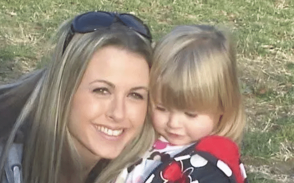 A smiling woman with long blonde hair and sunglasses on her head poses closely next to a young blonde girl wearing a colorful polka-dotted jacket outdoors.