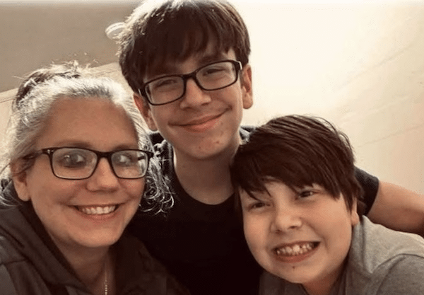A woman with glasses smiles for a selfie with two smiling boys, one of whom is wearing glasses, all posing closely together indoors.