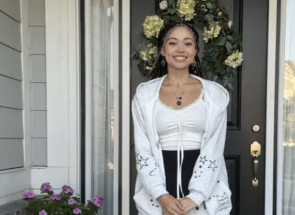 A young woman with braided hair stands smiling in front of a black door adorned with a floral wreath, wearing a white top, white jacket with star patterns, and a black skirt; a potted plant with pink flowers sits to her left.