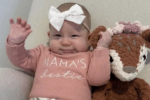A smiling baby wearing a large white bow headband and a pink outfit that says "Mama's bestie," sitting on a couch next to a stuffed animal.