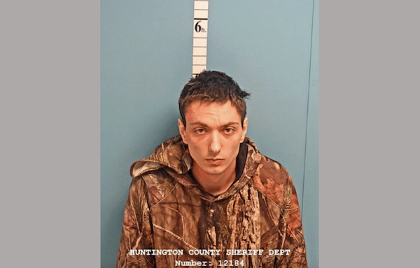 A young man wearing a camouflage hoodie stands in front of a height measurement wall, looking straight at the camera.