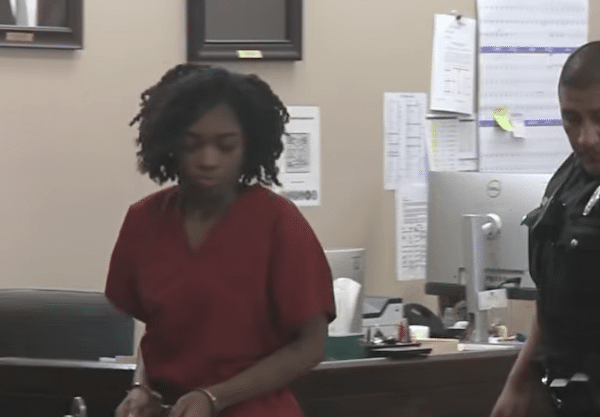A woman in a red jumpsuit stands beside a uniformed officer inside a courtroom, with framed photos on the wall behind them.