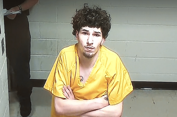 A man with curly hair wearing a yellow prison uniform stands with his arms crossed in a jail holding area, looking away from the camera.