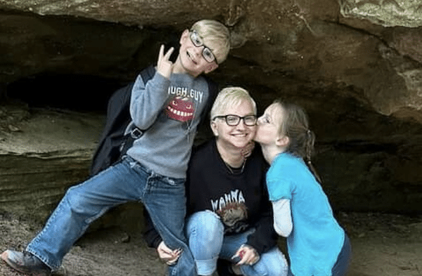 A woman with short blonde hair and glasses kneels outdoors in front of a rock formation, smiling as a young girl kisses her cheek and a boy stands on her other side, making a peace sign with his hand.