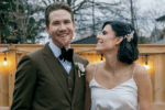 A smiling bride and groom pose together outdoors, the groom in a brown suit with a boutonnière and the bride in a white dress with a floral hair accessory, standing in front of a wooden fence with string lights.