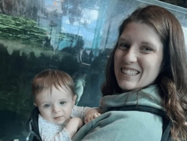 Woman with brown hair smiling in front of a glass aquarium, partially visible child next to her.