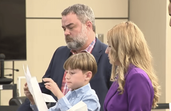 A young boy reads from a sheet of paper while a bearded man in a blazer and a woman in a purple jacket stand beside him in a courtroom.