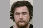 Mugshot-style image of a man with dark wavy hair and facial hair, facing the camera with both eyes closed or missing.