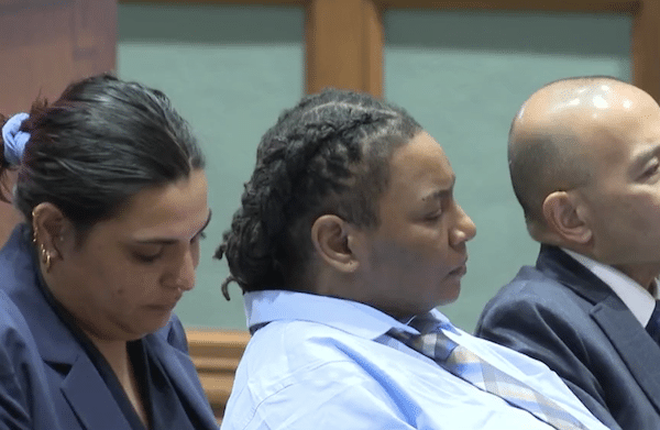Three people sit side by side in a courtroom, with the central person in a light blue shirt and striped tie shown in profile.