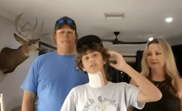 A teenage boy with a bandage on his neck stands between his parents inside a home, touching his hair while the adults look on.