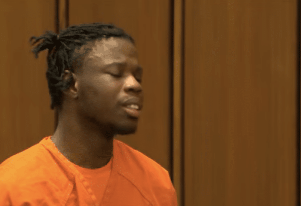 A man in an orange jail uniform stands in a courtroom with his eyes closed against a wood-paneled background.