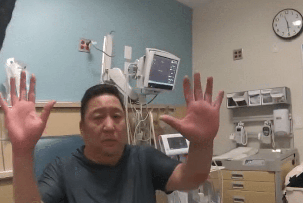 Man raises both hands while sitting in a hospital exam room with medical monitors behind him.