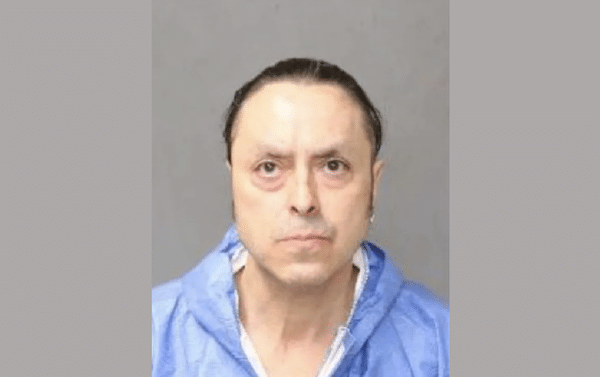 Mugshot-style image of a man with dark hair pulled back, wearing a blue jail uniform, facing the camera against a gray background.