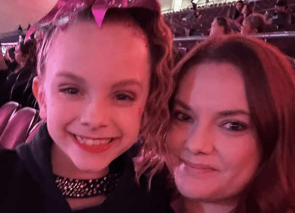 A smiling young girl with a pink bow and cheer-style hairstyle poses beside an adult woman as they take a selfie in arena seating under pink lighting.