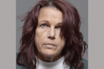 Close-up booking photo of a woman with shoulder-length dark red hair, wearing a white high-neck top and dark outer layer, looking straight at the camera.