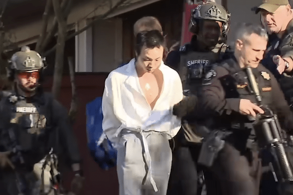 Sheriff’s deputies and tactical officers escort a handcuffed man in a white shirt outside a building during an arrest.