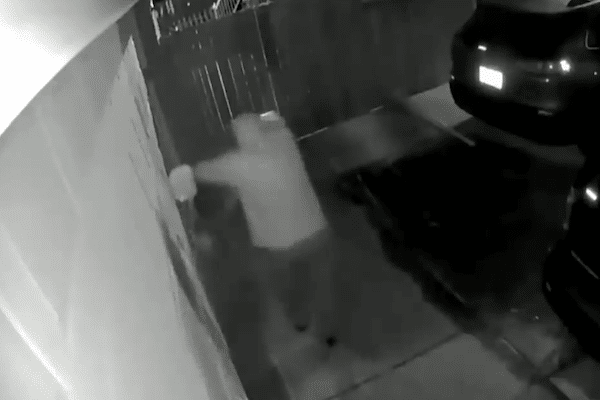 Black-and-white surveillance footage shows a hooded person crouching near the side of a house at night beside a parked SUV in a driveway.