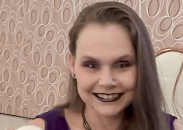 Woman with long brown hair wearing dark lipstick and purple eye makeup smiles in front of patterned wallpaper.