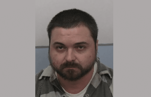 Mugshot-style photo of a bearded man with short dark hair wearing a gray striped jail uniform, facing the camera with a neutral expression.