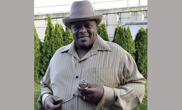 Man standing outdoors in a tan pinstriped shirt and matching hat, holding a pocket watch chain and smiling at the camera.