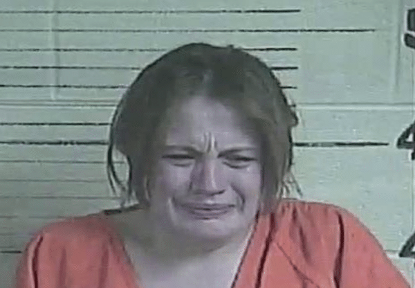 A woman wearing an orange jail uniform appears to be crying during a booking photo, with a lined height chart visible behind her.