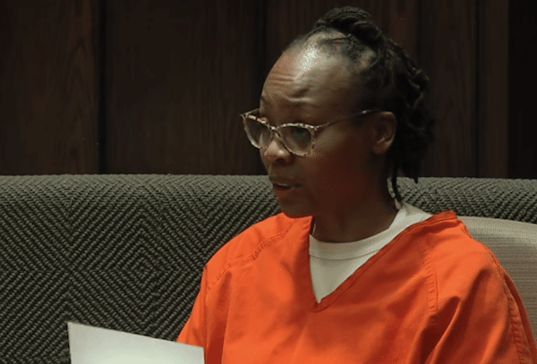 A woman in an orange jail uniform and glasses sits in a courtroom chair, looking down at papers in her hands.