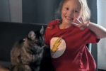 Smiling young girl in a red Flash T-shirt poses with one hand raised beside a fluffy tabby cat on a dark chair.