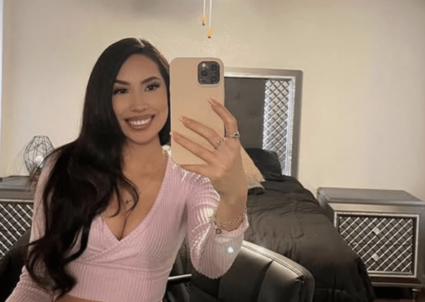 Smiling woman taking a mirror selfie in a bedroom, wearing a light pink long-sleeve wrap crop top and jeans, with a bed and mirrored nightstands in the background.