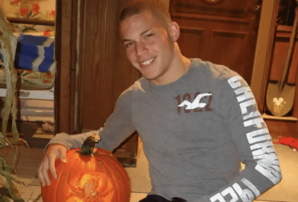 A young man in a gray long-sleeve shirt with "California" written on the sleeve sits on a porch, smiling at the camera next to a carved and lit pumpkin.