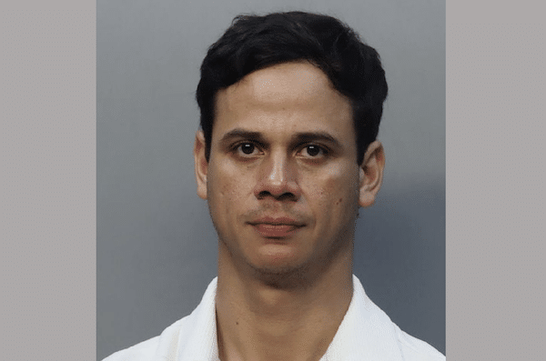 Mugshot-style head-and-shoulders photo of a dark-haired man in a white collared shirt against a gray background.