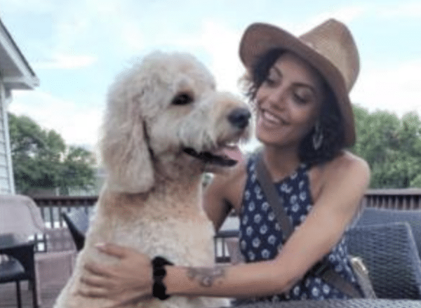 A woman in a brown hat hugs a large curly-haired dog while smiling on an outdoor deck.